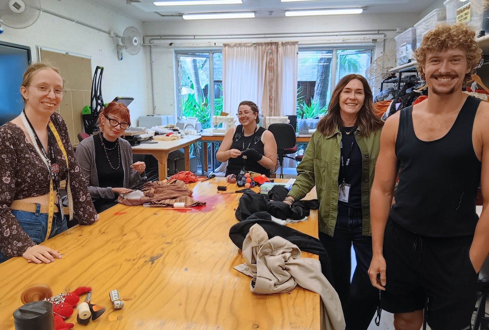 A group of five people stand smiling around a large wooden table filled with sewing materials and colourful fabric in a well-lit crafting room.