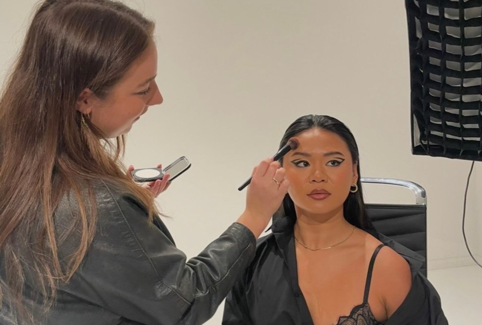 A make-up artist is applying make-up to a model in a black shirt which is falling off her shoulder.