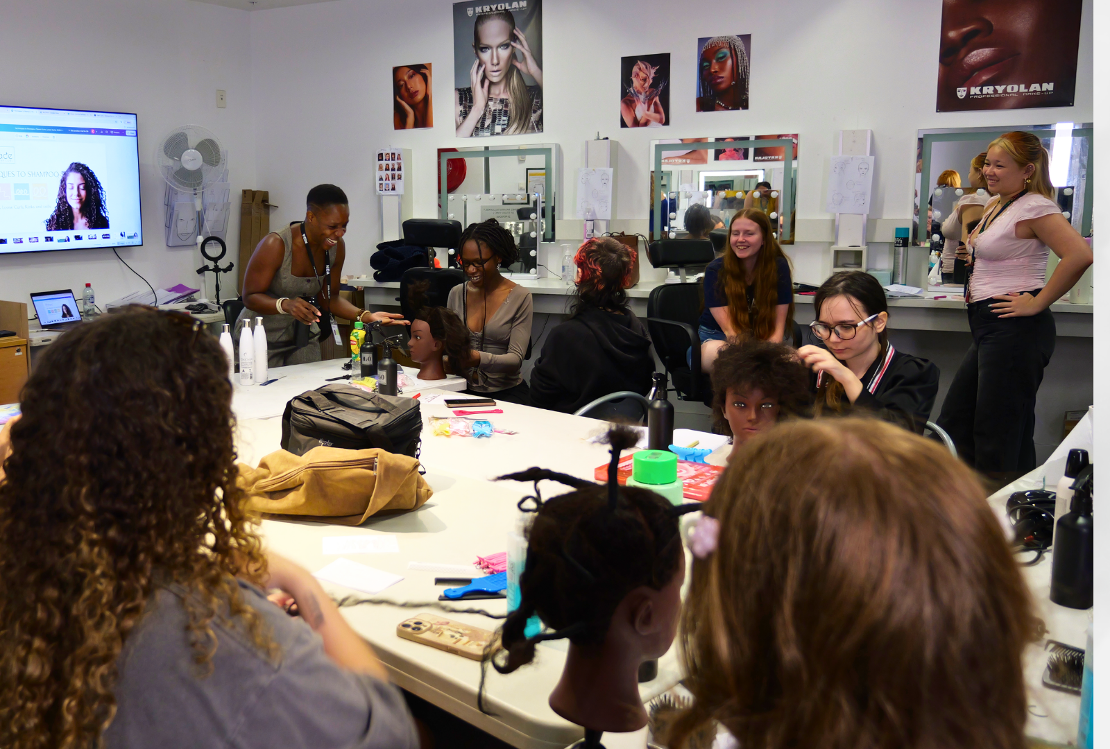 A group of students sitting around a table with mannequins styling their hair while a woman teaches. 