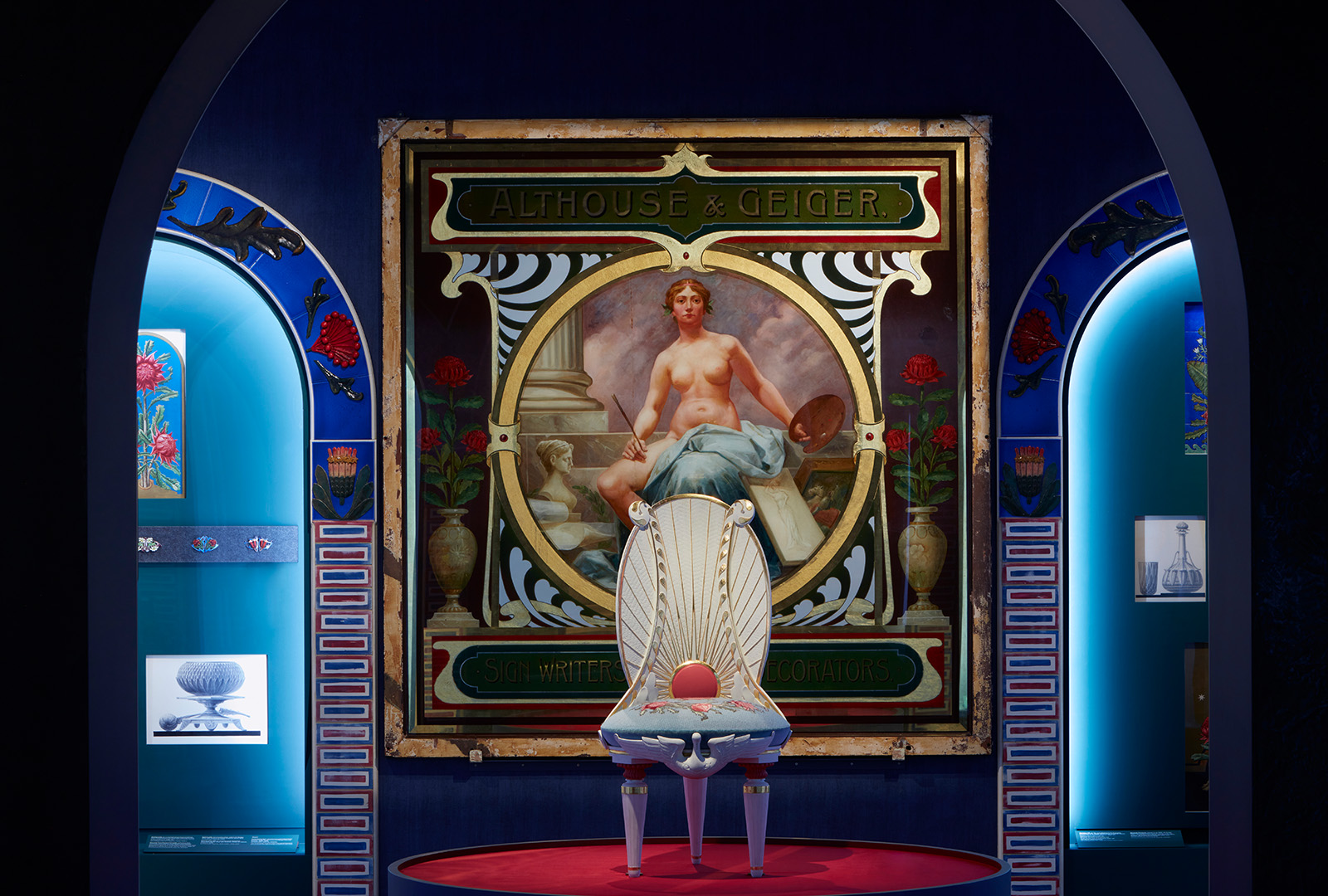A decorative display features a white ornate chair with a red cushion in front of a large vintage Althouse & Geiger poster depicting a seated woman. The setting is framed by arched alcoves with illuminated art pieces.