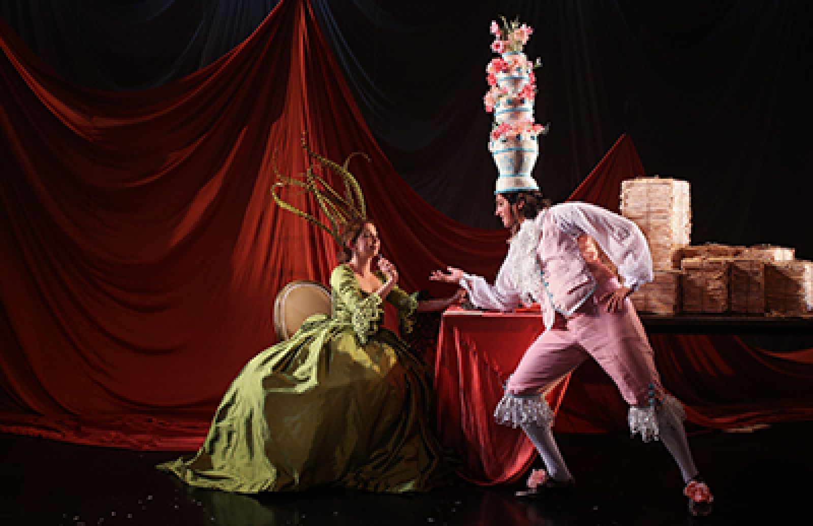 Two people in elaborate costumes perform theatrically. One, in a green dress and dramatic headpiece, sits on a chair; the other, in pastel pink with a tall cake hat, poses beside a table stacked with pancakes. Red drapes hang in the background.