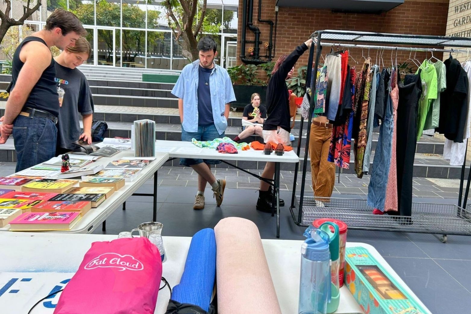 People shop at a colourful outdoor market. Tables display books, with clothing on racks. The mood is relaxed and communal.