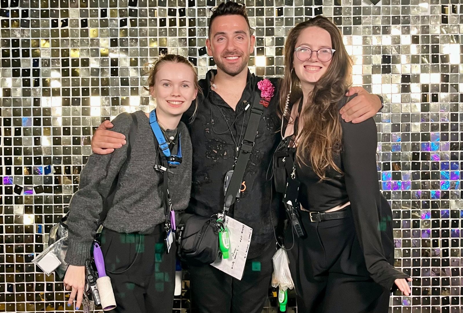 Three people smiling, standing close together against a shiny, sequined backdrop. They appear happy, dressed in dark attire, and wearing event lanyards.