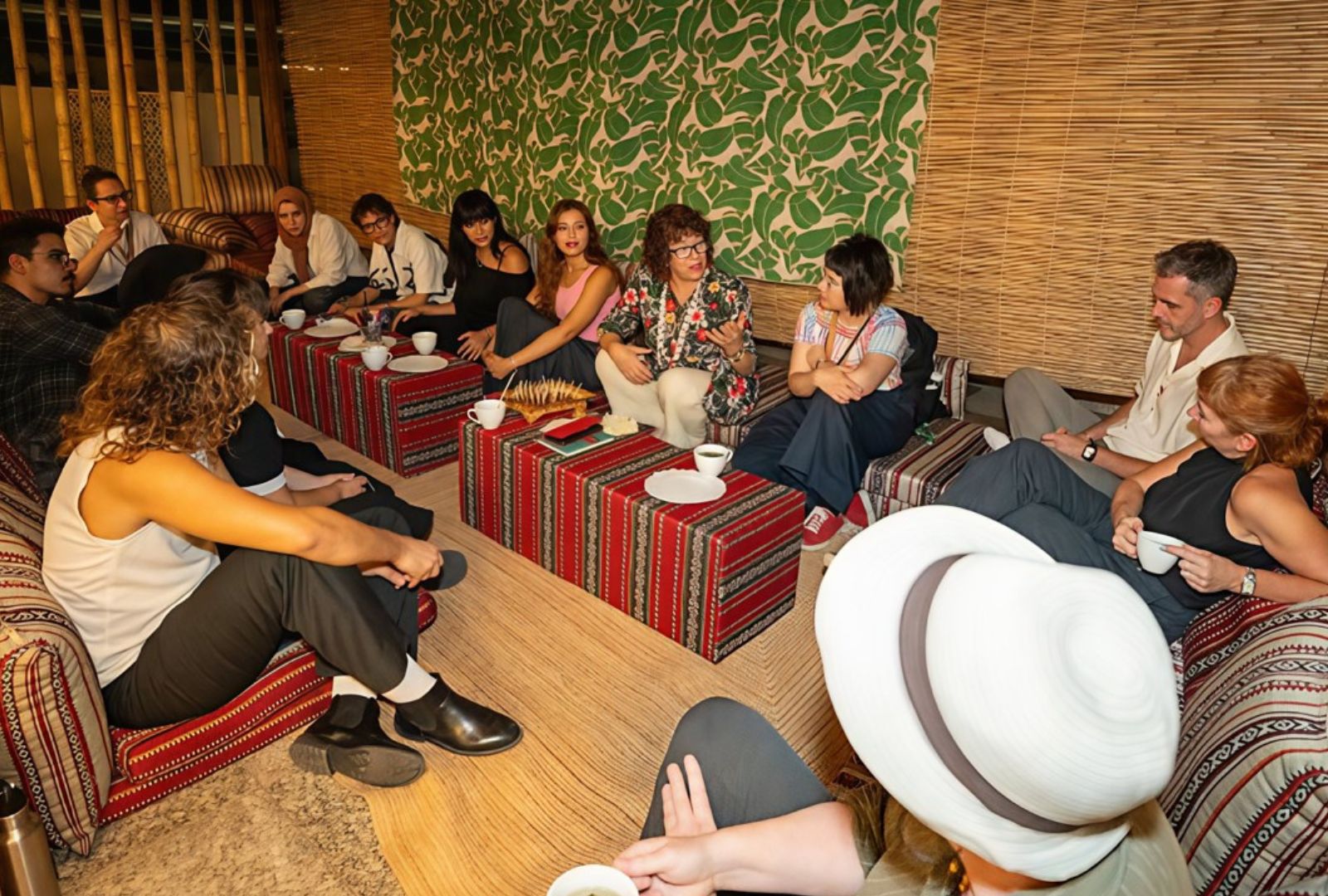 A diverse group of people sit in a circle on patterned cushions, engaging in a lively discussion. The setting is cosy, with a leafy wallpaper backdrop.