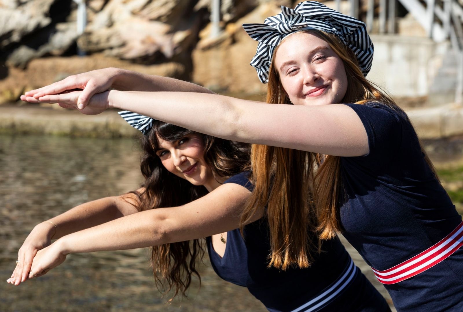 Two individuals wearing striped headbands and navy outfits pose in a playful, synchronised swimming gesture near water.