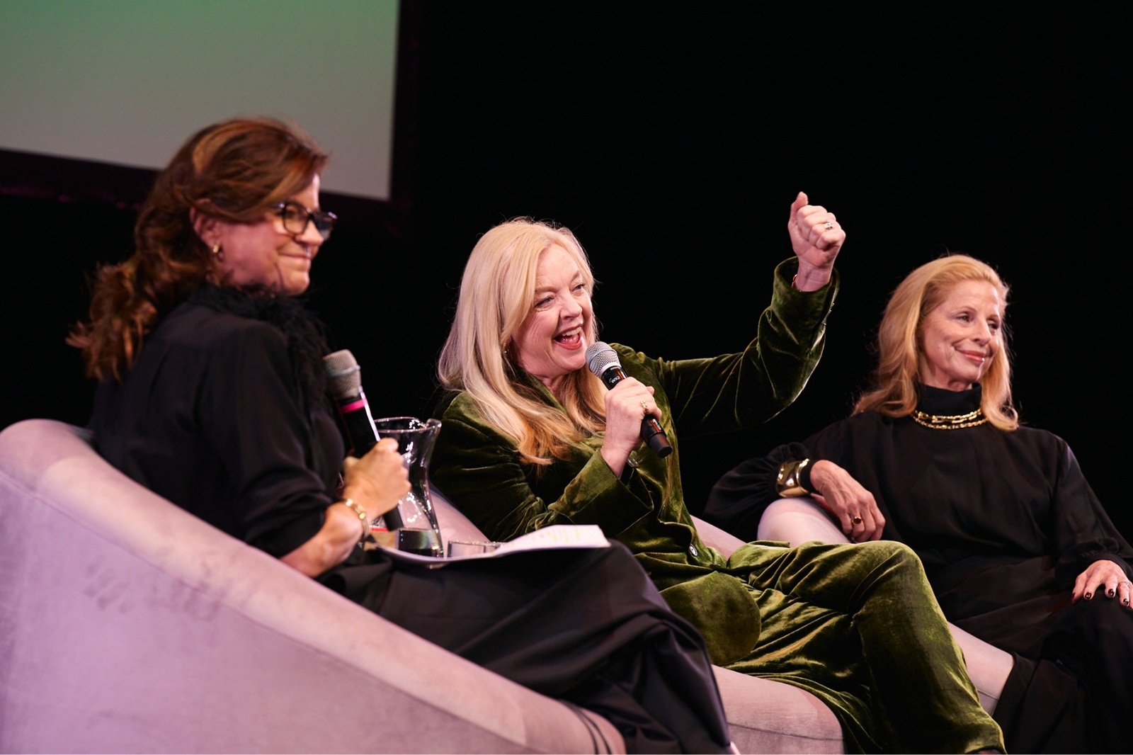 A woman in a green suit raises a fist in the air, holding a mic to her mouth signifying she is speaking to a crowd. Another woman smiles encouragingly behind her. 