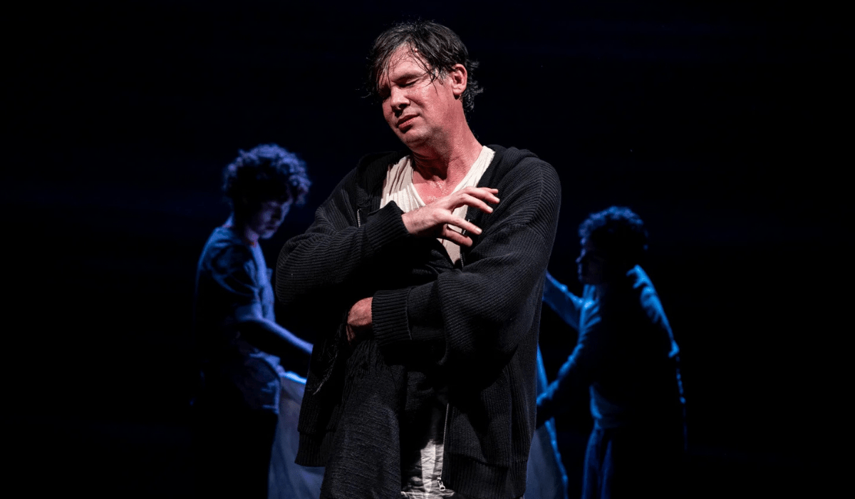 A man stands in dramatic stage lighting, eyes closed and hand on chest, conveying deep emotion. Two silhouettes are seen in the dark background.