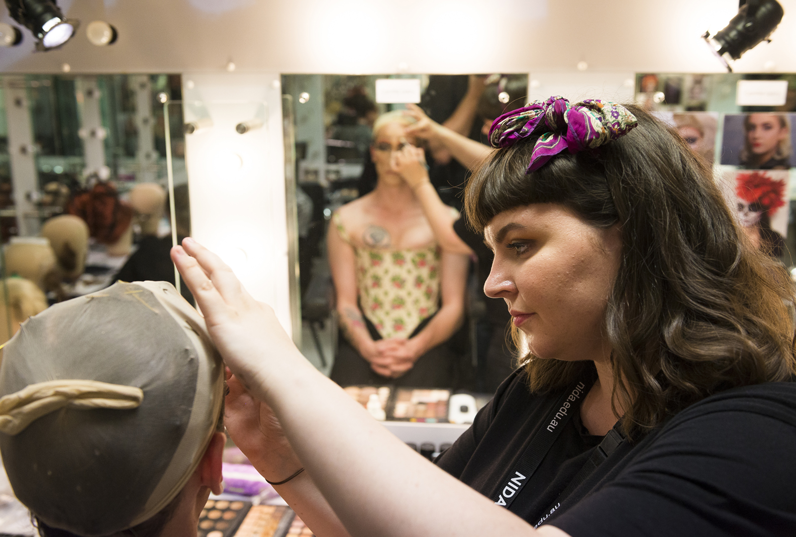 A make-up artist adjusts a wig cap on a person in a make-up room, while another person with floral face paint is reflected in the mirror in the background. Make-up tools and lights are visible on the dressing table.