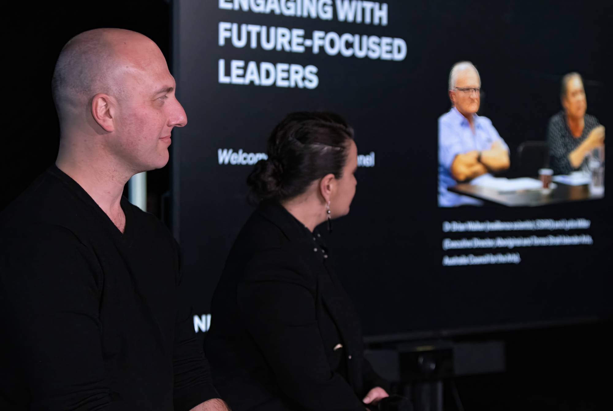 Two people sit side by side in front of a screen displaying a presentation titled Engaging with Future-Focused Leaders, showing two more people speaking in a video call. The setting appears to be a panel discussion or seminar.