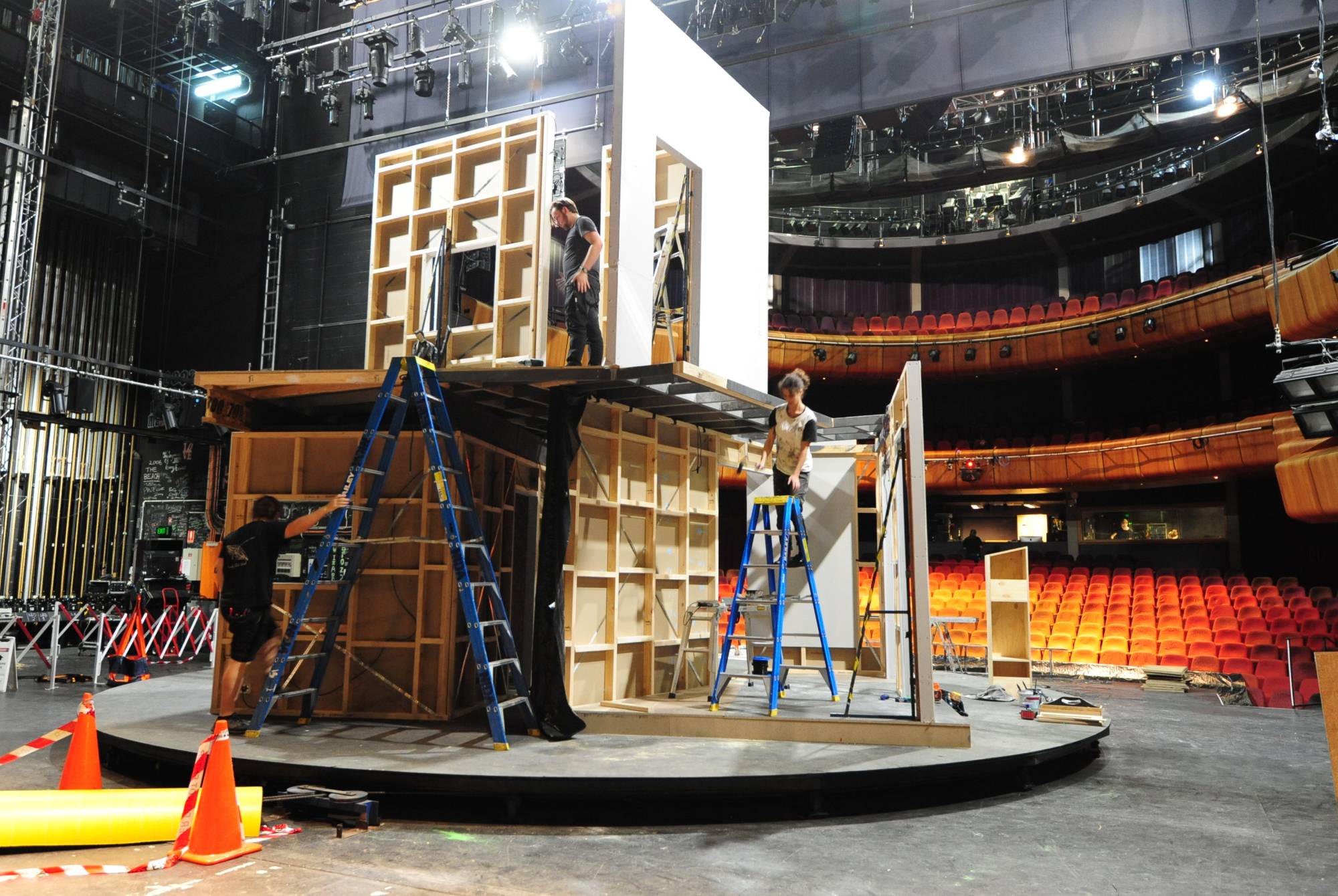Several people stand on ladders assembling a large stage set with wooden frames in a theatre, surrounded by empty red seats and stage equipment.