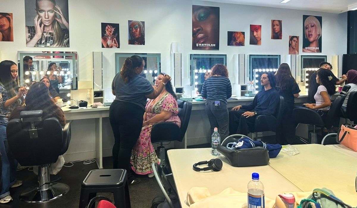 A makeup classroom with students applying makeup at mirrors. Posters of models line the walls. The atmosphere is lively and collaborative.