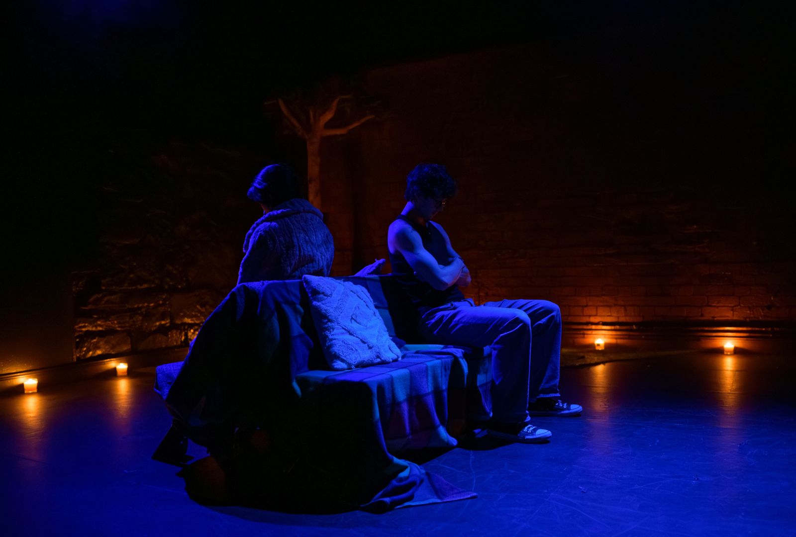 Two people sit back-to-back on a dimly lit stage with a dark, moody ambiance. Small candles line the floor, casting a soft glow.