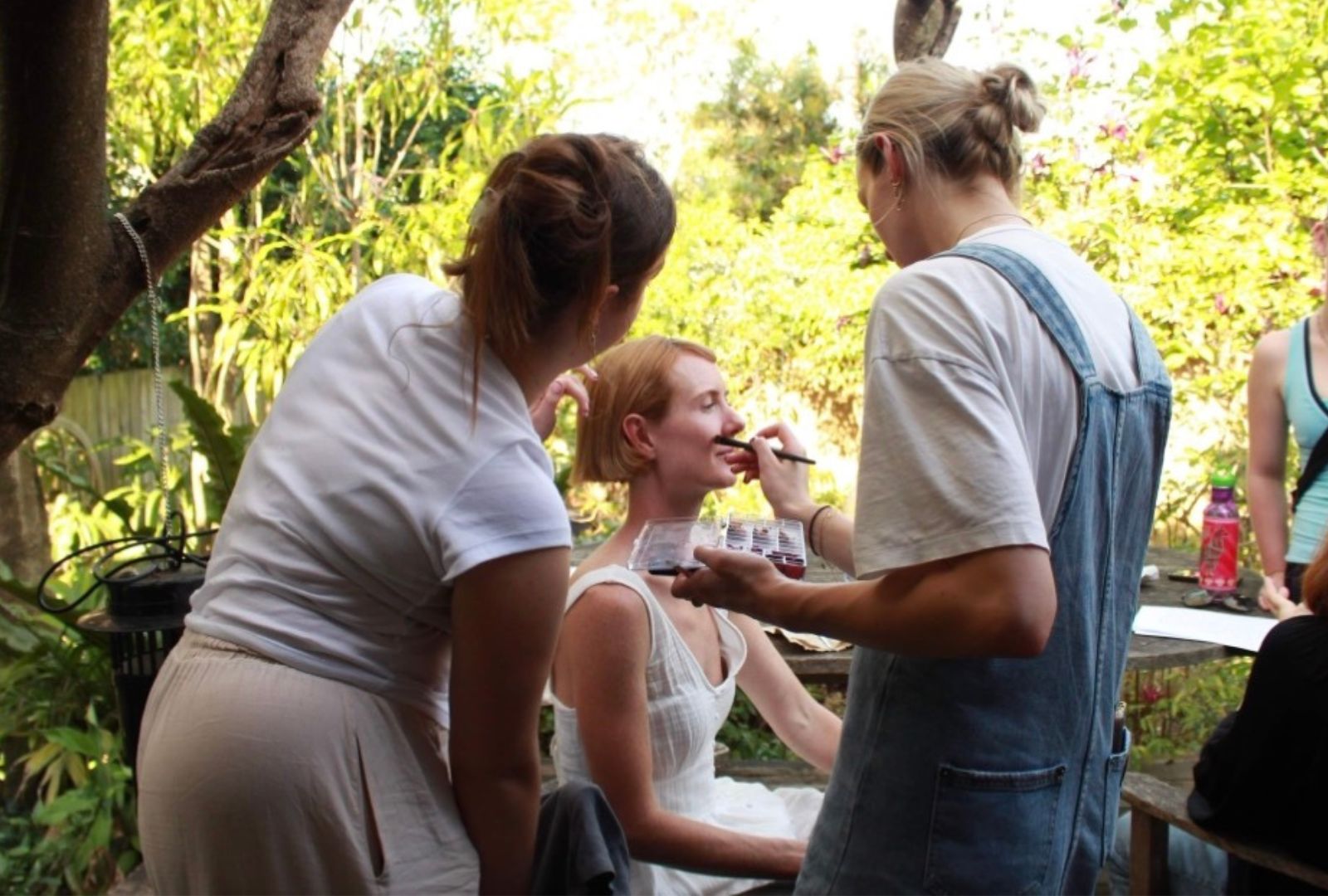 Two make-up artists are applying make-up to a model in a white dress outside.