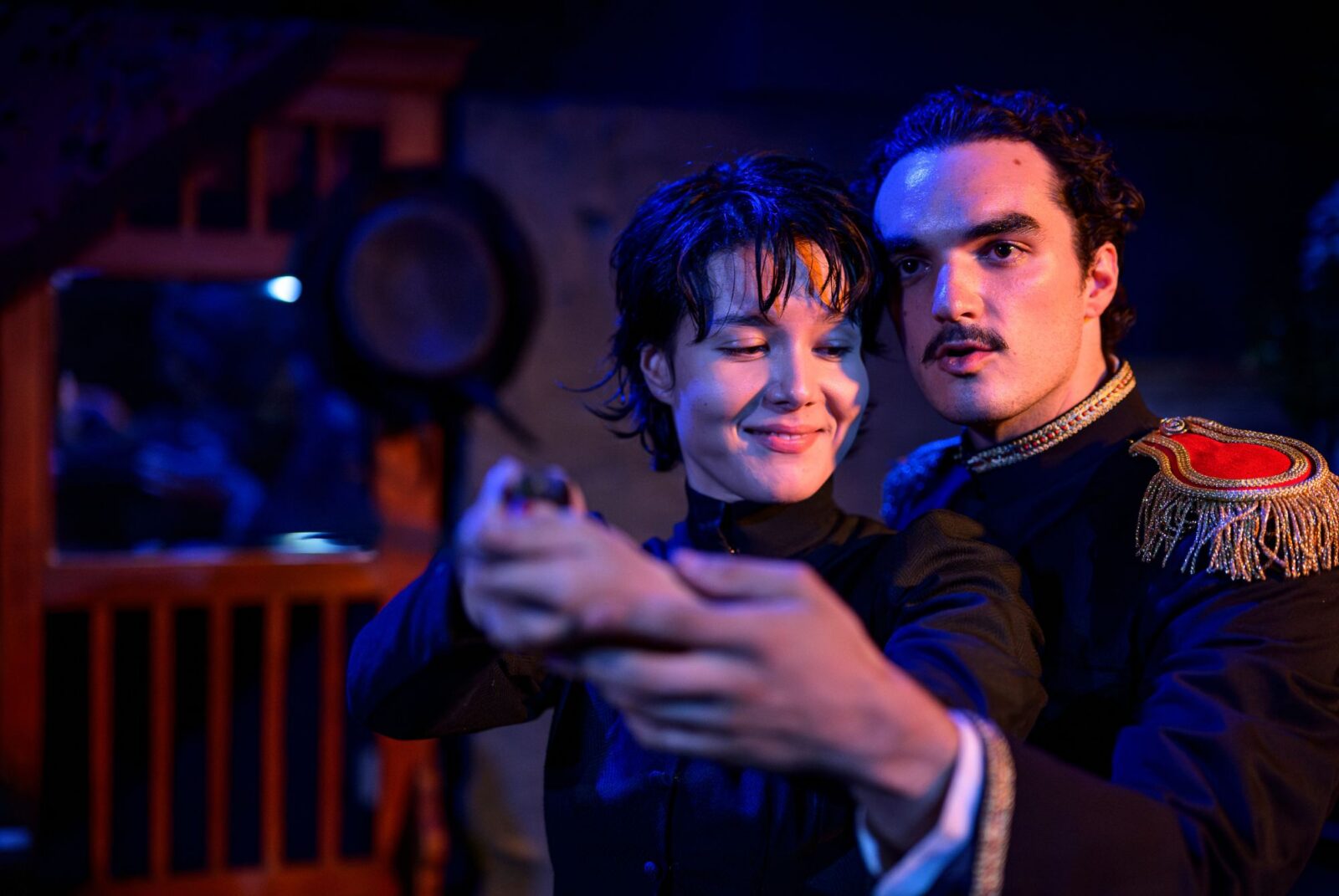 A person with short dark hair smiles while holding a gun, guided by another person in a military-style uniform. The scene is set in dim, dramatic lighting.