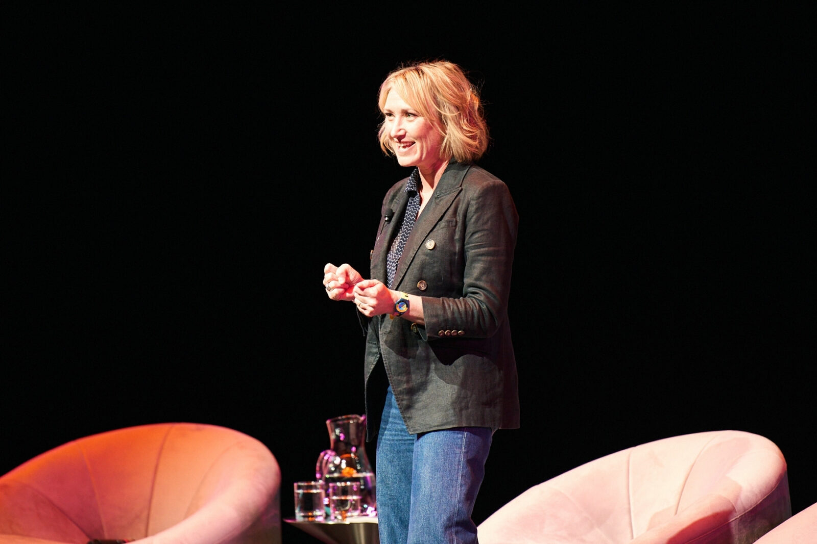 A woman in a black blazer speaks animatedly on  stage to a crowd. 