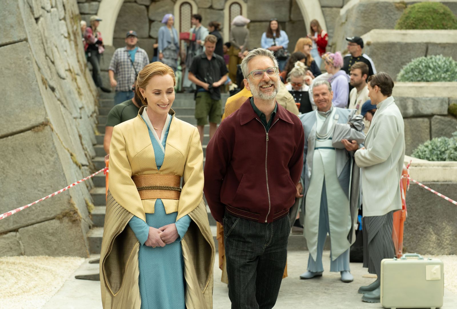 On set, an actor in a fantasy costume walks beside a costume designer with other crew and actors behind them. They are all smiling and look happy.