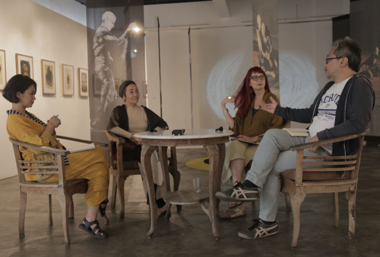Four people sit around a round table in an art gallery, engaged in conversation. Artworks hang on the walls behind them.