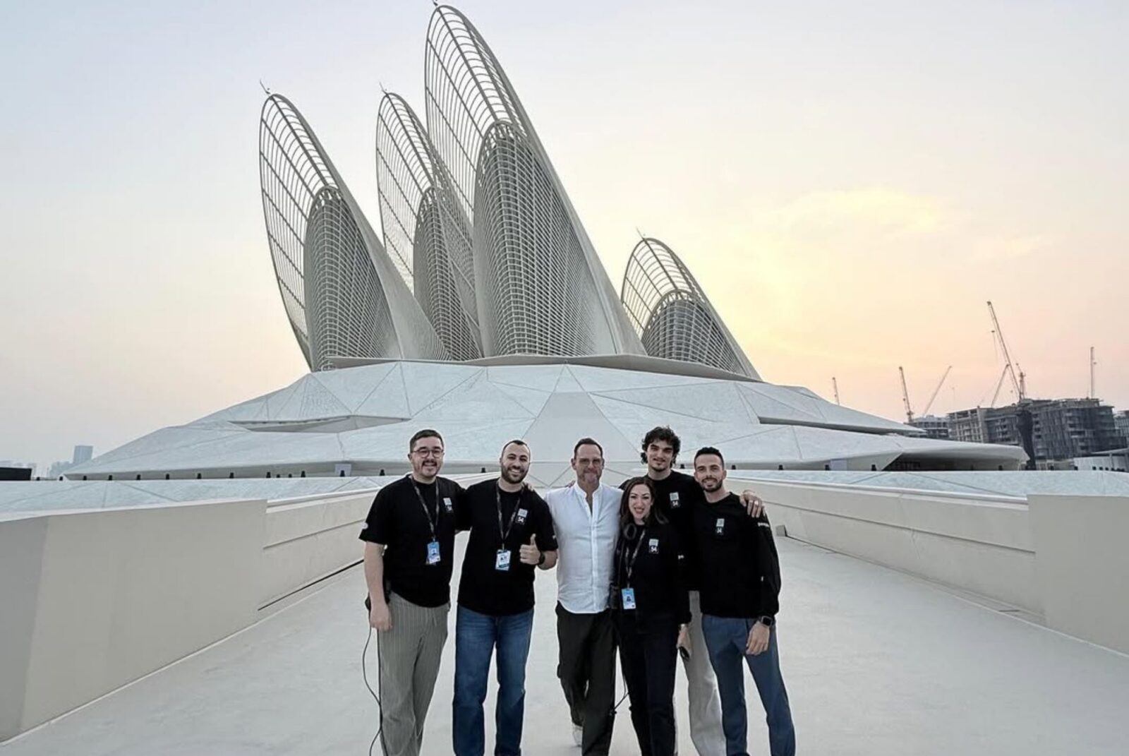 A group of six people, smiling and standing close together, in front of a futuristic building with several large, overlapping, sail-like structures at sunset.