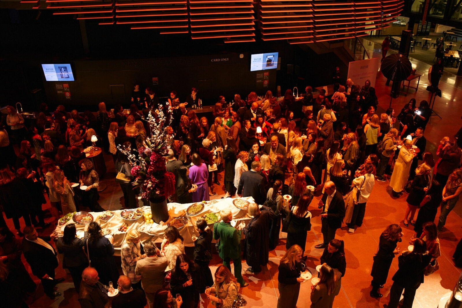 A large crowd of people attending an event at a venue with banners from NIDA hanging above. The space is warmly lit, and guests are mingling around a central table with refreshments.