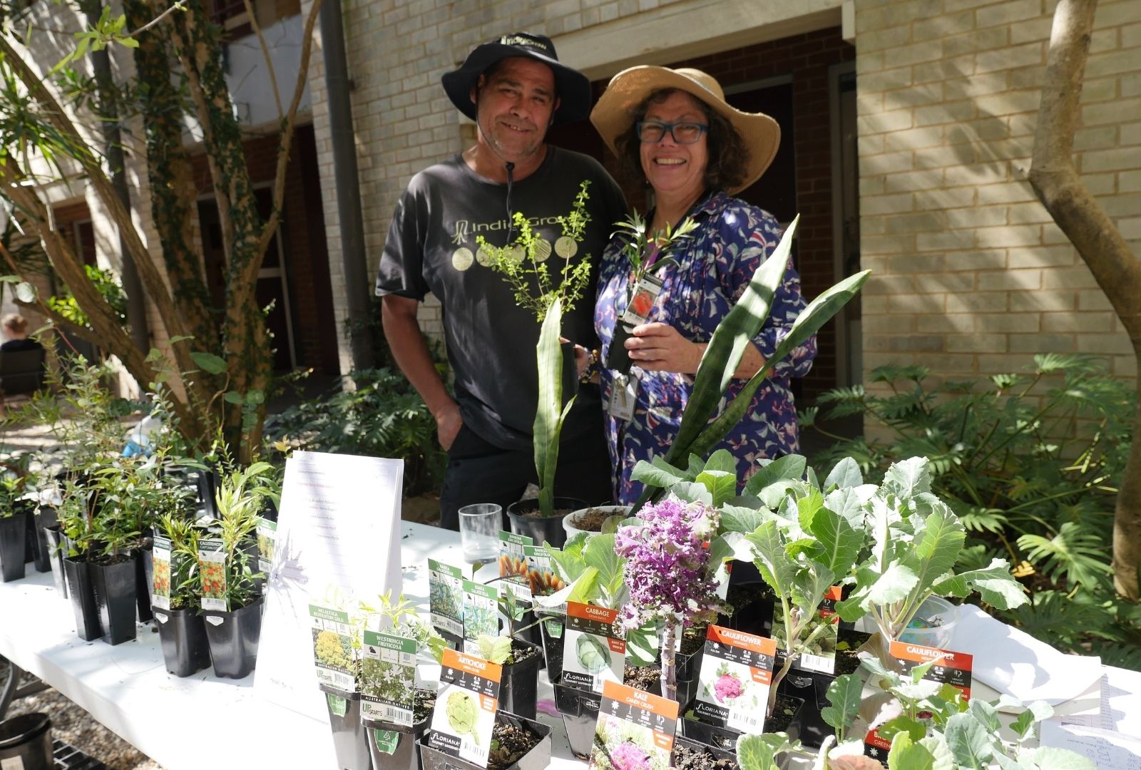 A smiling man and woman in sun hats stand behind a table filled with potted plants and seedlings, outdoors on a sunny day, exuding a cheerful, garden-themed atmosphere.
