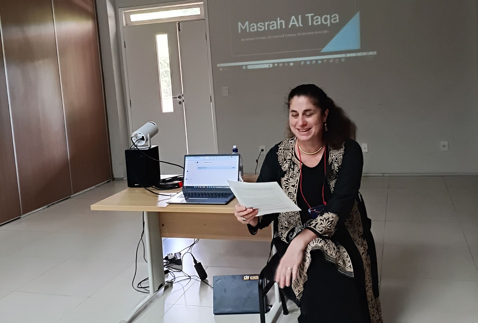 A person sitting beside a table presenting work. On the wall it reads 'Masrah Al Taqa'.