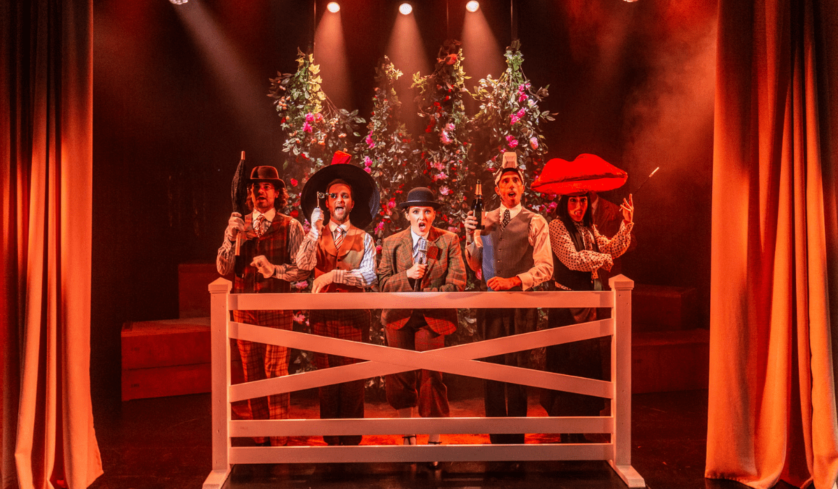 Five people in colourful costumes and hats stand behind a white fence on a stage decorated with greenery and flowers, with spotlights shining above.