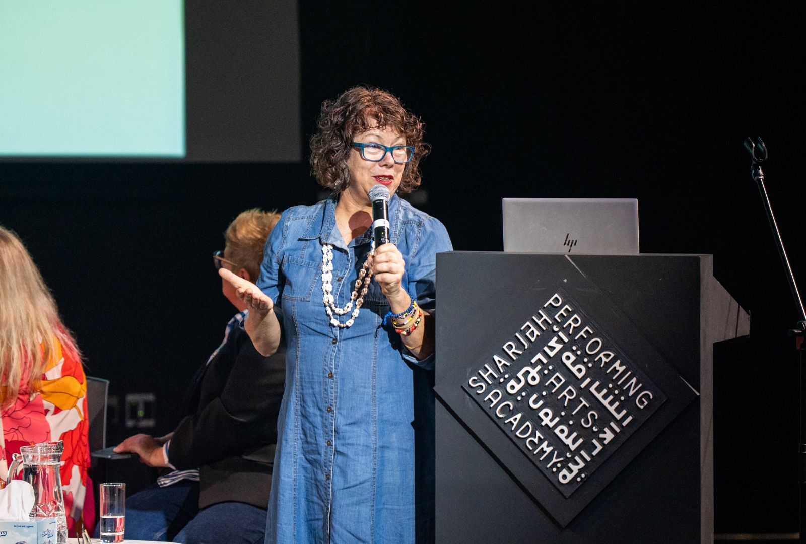 A woman in blue glasses speaks into a microphone at a podium with "Performing Arts Academy." She's wearing a blue dress and colourful accessories.