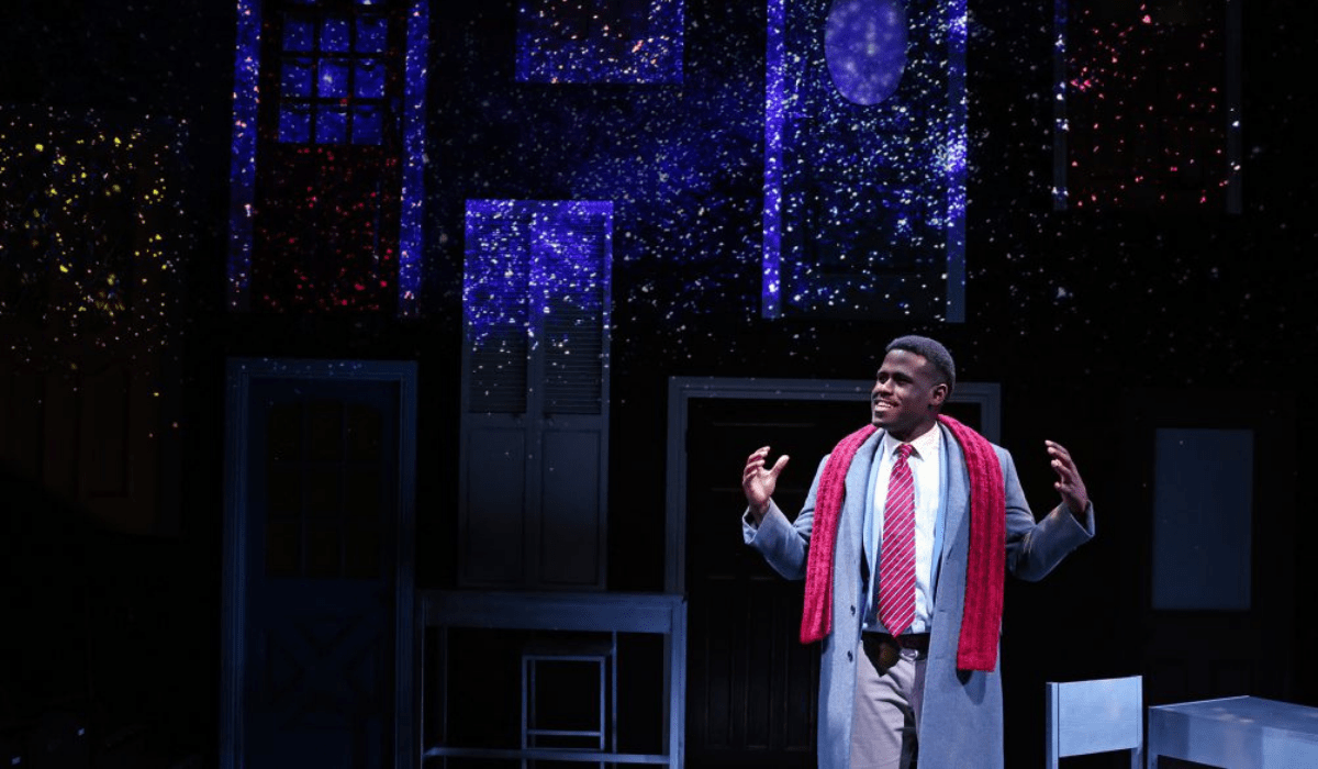 A man in a grey coat and red scarf stands on a dimly lit stage, surrounded by glowing stars. He appears thoughtful, set against a backdrop of windows.