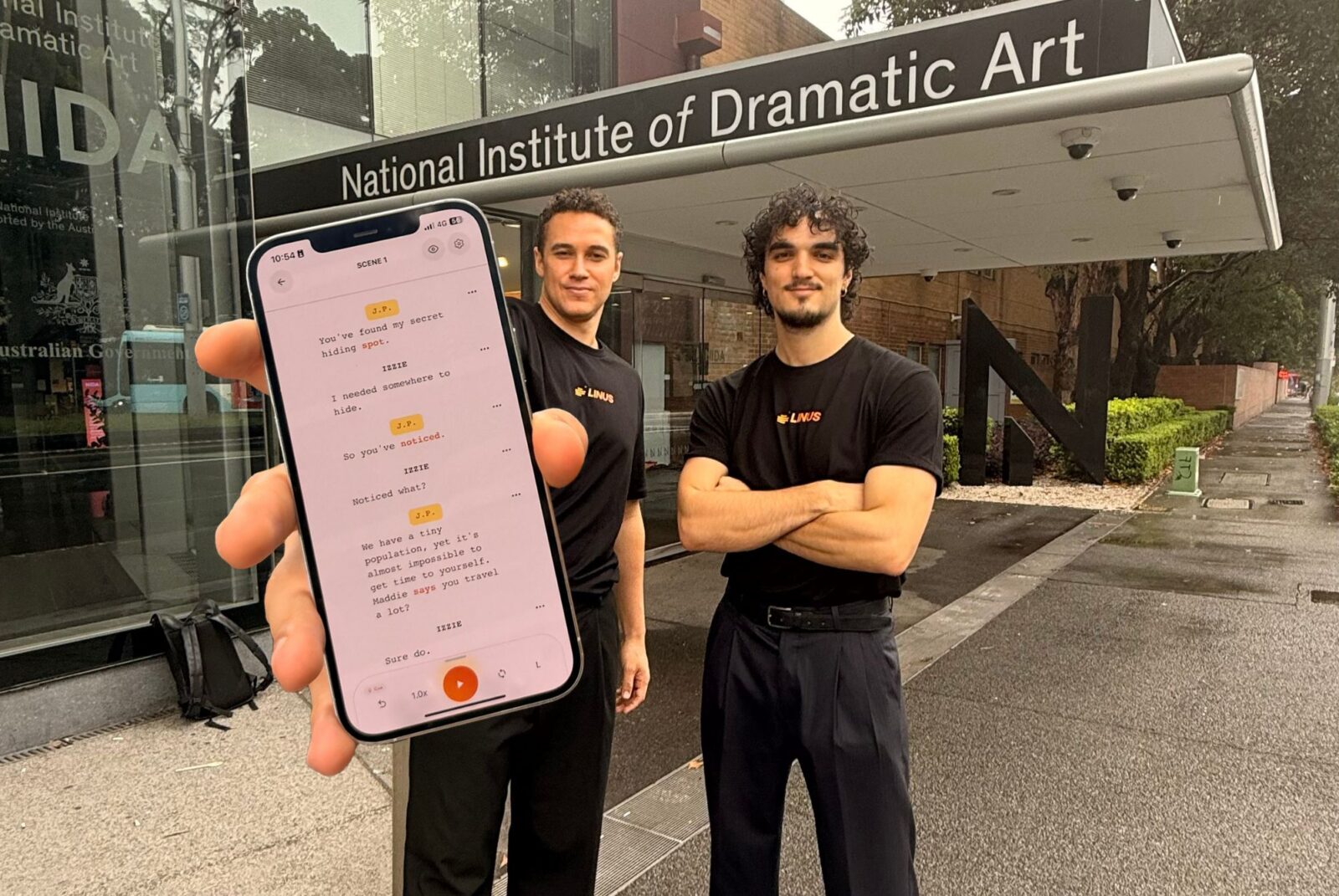 two men stand in front of the National Institute of Dramatic Art. One has his arms crossed, the other holds his phone out to the camera.
