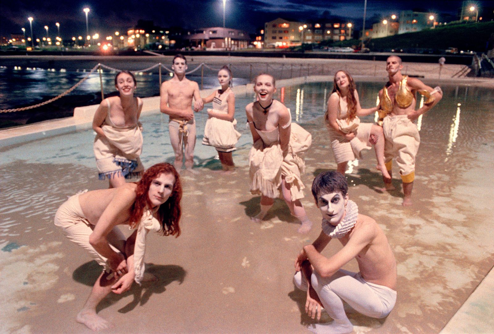 A group of eight people with painted faces and theatrical costumes pose playfully in a shallow, illuminated pool at night, exuding a whimsical, artistic vibe.