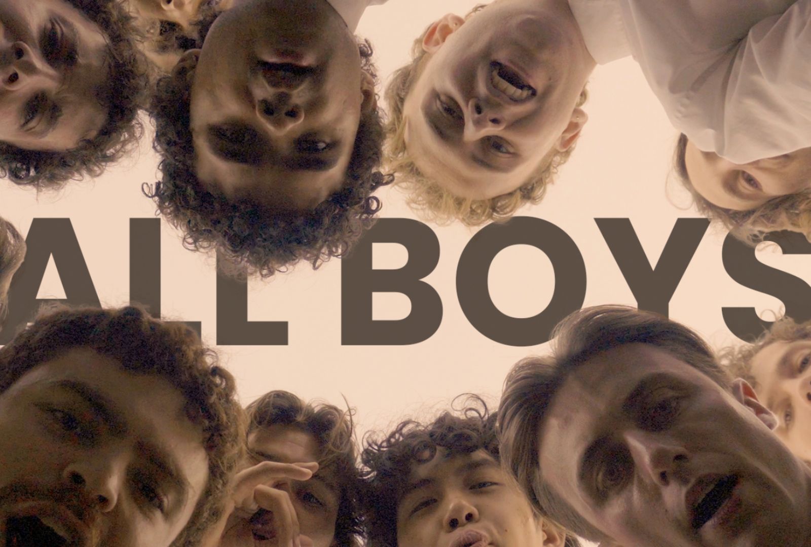 The cast of ALL BOYS crowd around a camera so only their faces are visible, in the background 'ALL BOYS' is written in brown lettering.