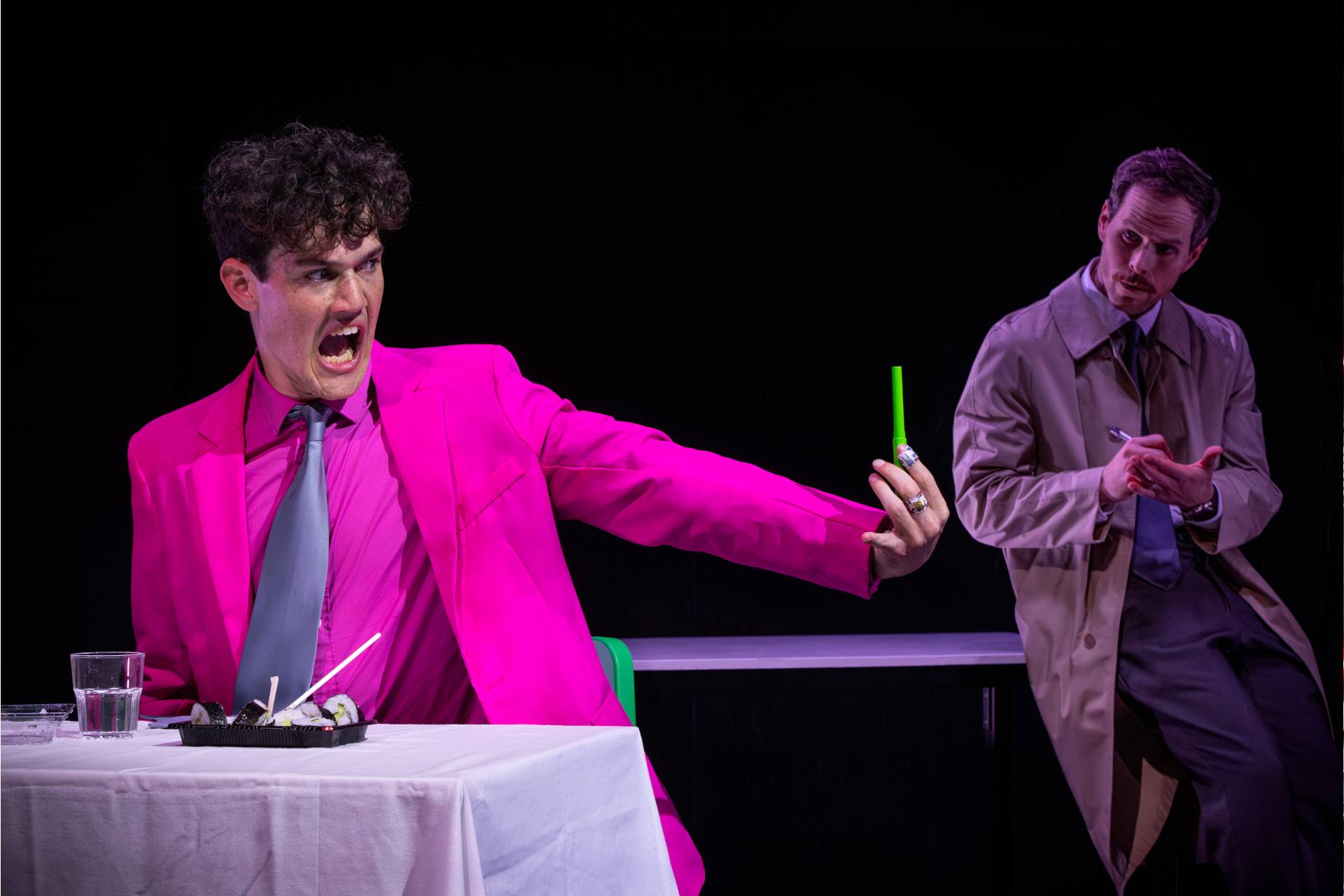 A man in a bright pink suit angrily holds up a green object at a table with food, whilst another man in a beige trench coat looks on sceptically in the background.