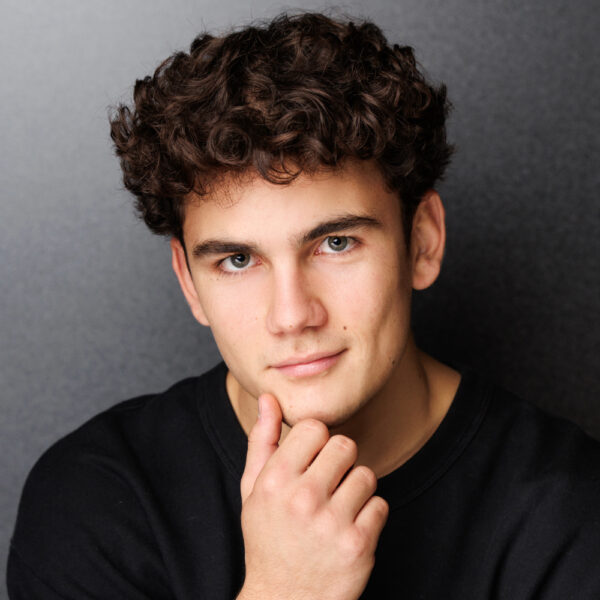 A young man with curly dark hair and light skin looks at the camera, resting his chin on his hand and wearing a black shirt, with a neutral dark background.
