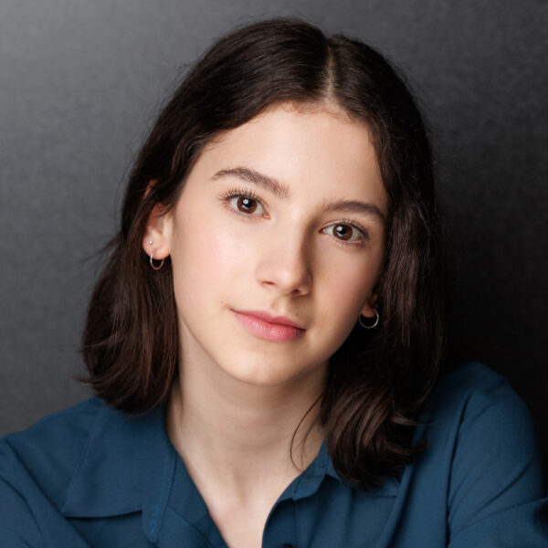 A young person with shoulder-length dark brown hair, wearing a blue shirt and small hoop earrings, looks directly at the camera with a neutral expression against a dark background.