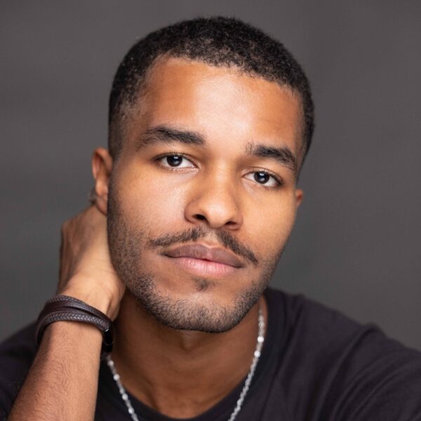 A young man with short hair and a trimmed beard looks at the camera, wearing a black shirt and a necklace, with one hand resting behind his neck. The background is plain and neutral.