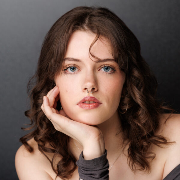 A young woman with wavy brown hair and blue eyes rests her chin on her hand, gazing at the camera. She has a neutral expression and wears a dark sleeve on her wrist. The background is plain and dark.