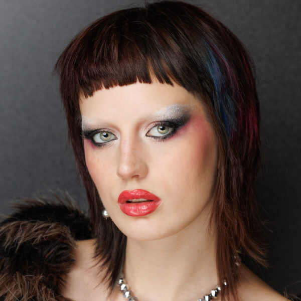 A person with a short, edgy haircut wearing bold silver and black eye make-up, red lipstick, and a fur-trimmed garment poses against a dark background. They have fair skin and intense eye contact with the camera.