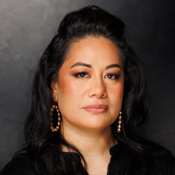 A woman with long, dark hair and hoop earrings, wearing a black top, gazes calmly at the camera against a dark, neutral background.