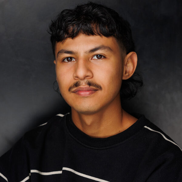 A young man with short dark hair, a moustache, and a faint beard looks at the camera. He wears a black jumper with white stripes and sits against a dark, simple background.