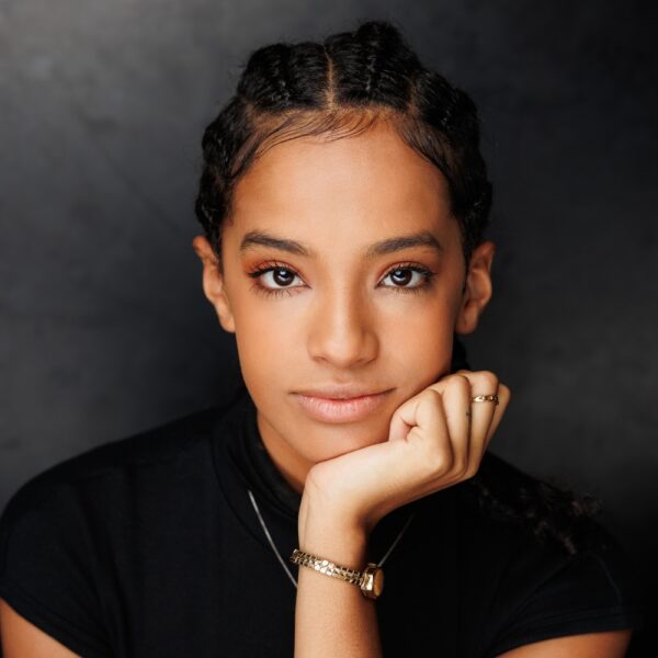 A young woman with plaited hair rests her chin on her hand, looking directly at the camera. She wears a black top, gold bracelet, and ring, with a calm, confident expression against a dark background.
