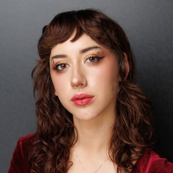 A young woman with wavy brown hair, winged eyeliner, and neutral make-up. She has facial piercings on her nose, eyebrow, and lower lip, and is wearing a burgundy top with a subtle necklace against a grey background.