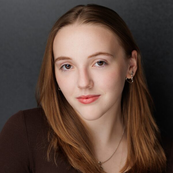 A young woman with straight light brown hair and fair skin is wearing a brown top, silver hoop earrings, and a necklace. She is looking at the camera with a slight smile against a dark, plain background.