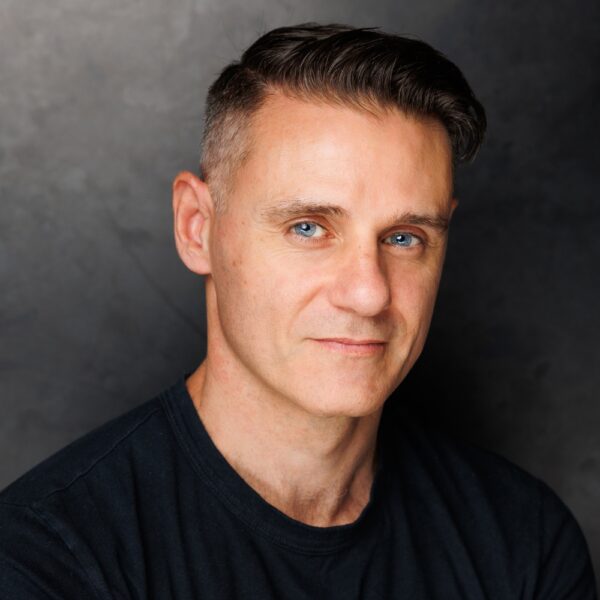 A man with short brown hair and blue eyes wearing a black shirt, looking at the camera with a slight smile against a dark grey background.