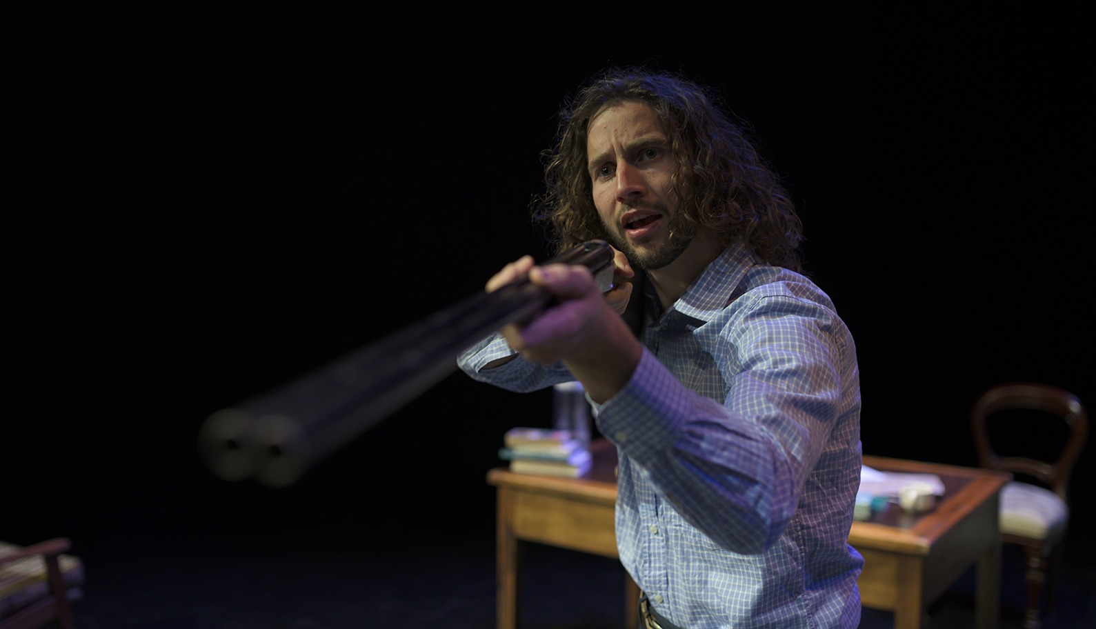 A man with long curly hair and a checked shirt points a double-barrelled shotgun towards the camera, looking intense, with a desk and chair in the background on a dark stage.