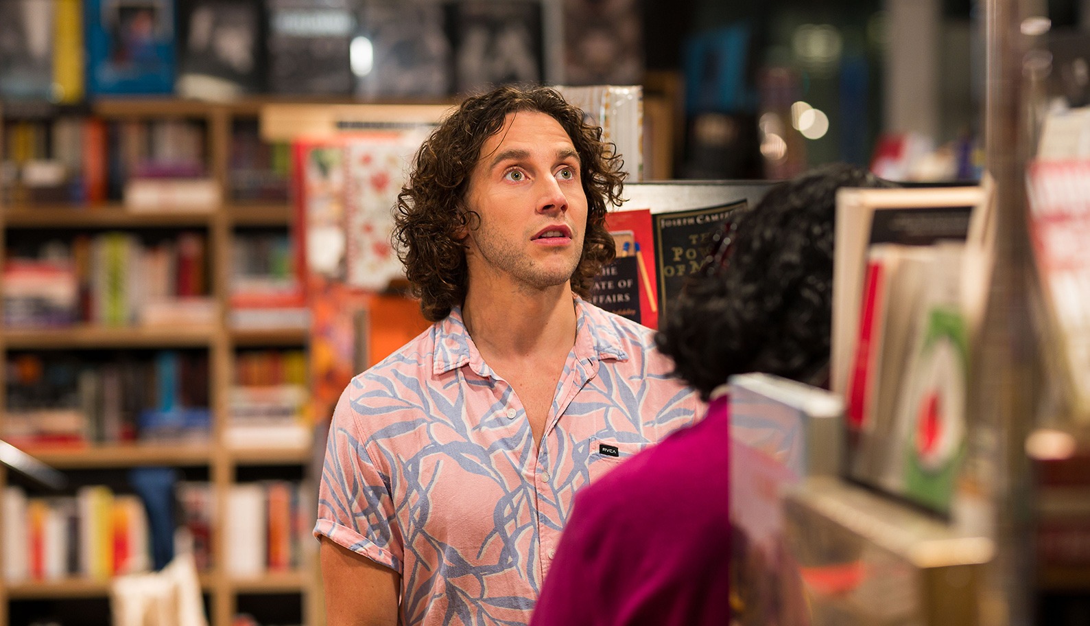 A man with curly hair wearing a pink patterned shirt stands in a bookshop, looking up with a surprised expression. Bookshelves and blurred books are visible in the background.