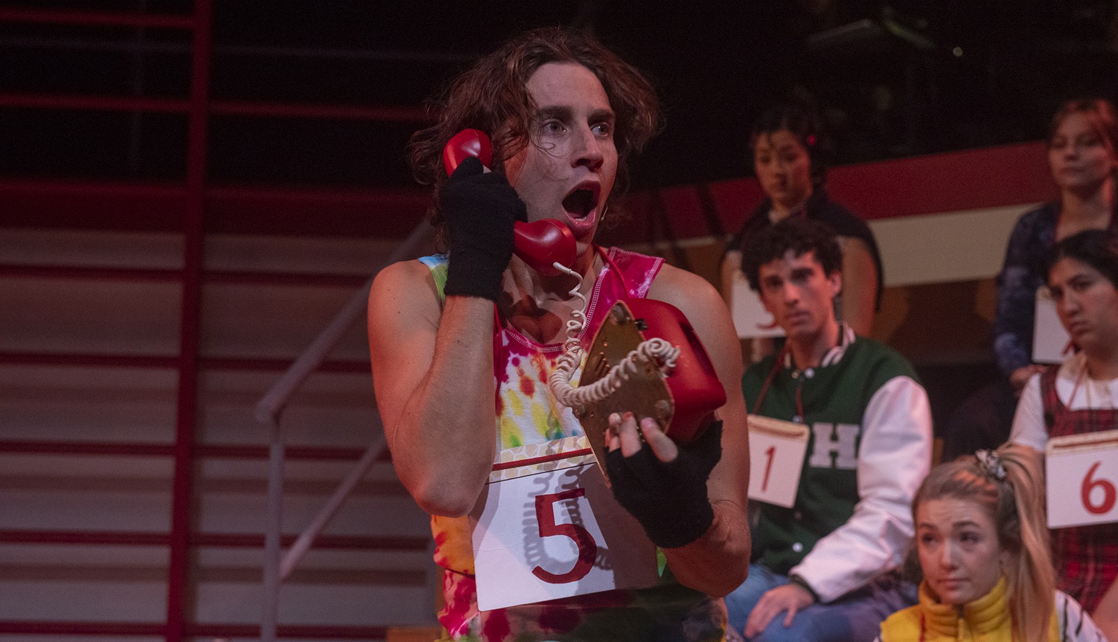 A person wearing a tie-dye shirt and black gloves holds a red rotary phone to their ear, looking surprised. Several people with numbered signs sit in the background, as if in a competition or spelling bee.