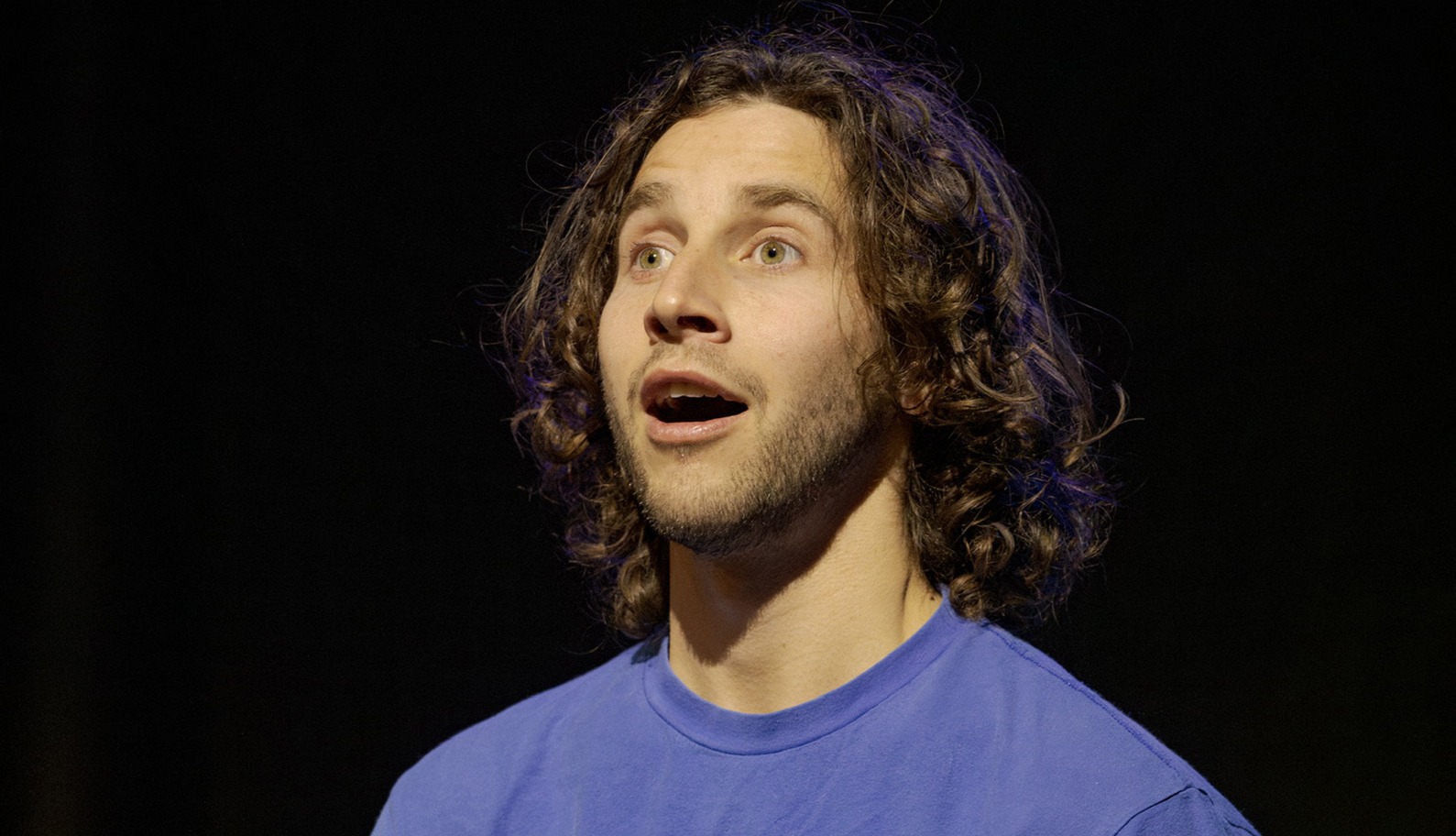 A man with long curly hair and a beard, wearing a blue shirt, looks upwards with a surprised or awed expression against a dark background.