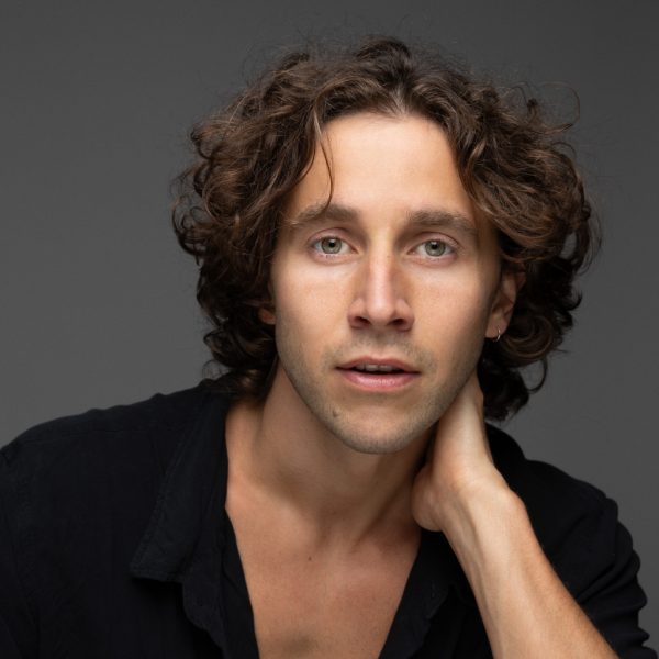 A man with wavy brown hair and green eyes gazes at the camera, wearing a black shirt with an open collar. He has his right hand resting on the side of his neck against a neutral grey background.