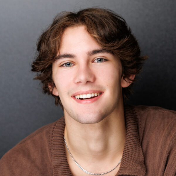 A young person with medium-length brown hair and blue eyes smiles at the camera. They are wearing a brown jumper and a silver chain necklace, with a dark grey background behind them.