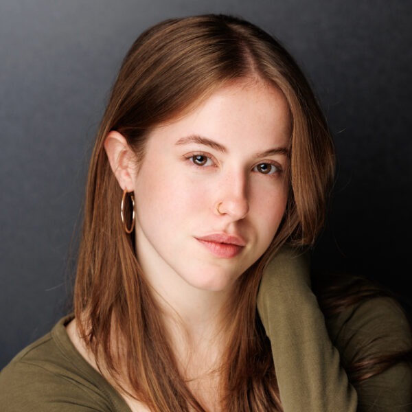 A young woman with long brown hair wearing a green top, hoop earrings, and a nose ring, gazes softly at the camera against a dark, plain background.